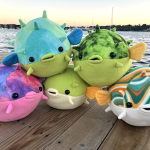 May include: A collection of five plush pufferfish toys featuring various patterns and colours, including tie-dye pink and blue, solid lime green, mottled dark green, and orange, green, and white striped designs, all with black eyes and soft spikes.