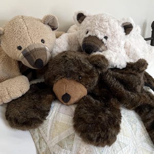 May include: Three plush teddy bears in shades of brown and white rest on a bed. The bears have soft fur, dark eyes, and plaid accents on their ears and noses. The bears are lying on a white and patterned quilt.