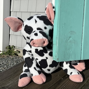 Könnte beinhalten: Ein schwarz-weißes Plüschtier in Form einer Kuh mit rosa Ohren und Hufen, das hinter einer türkisfarbenen Tür hervorlugt.