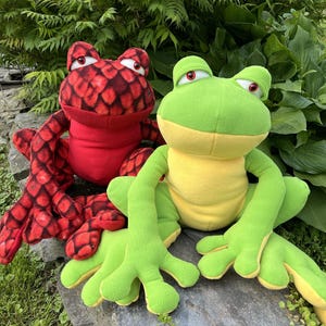 Peut inclure: Deux jouets en peluche en forme de grenouille. L'un est rouge avec un motif d'écailles, l'autre est vert avec un ventre jaune. Les deux ont de grands yeux et sont assis sur une pierre. Le fond présente un feuillage vert.