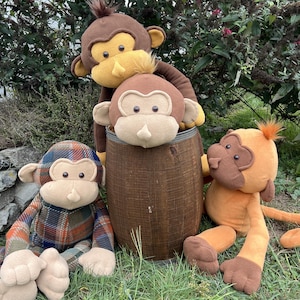 以下が含まれることがあります： 木製の樽の周りに配置された、茶色とオレンジ色のさまざまな色合いの4つのぬいぐるみ猿。 1匹の猿はチェック柄です。 猿はベージュの顔と耳をしており、一部には髪の房があります。