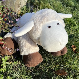 Könnte beinhalten: Ein weißes Plüschschafspielzeug mit braunen Hufen. Das Schaf hat ein flauschiges weißes Fell und braune Augen. Das Spielzeug liegt auf einem Bett aus grünem Gras.