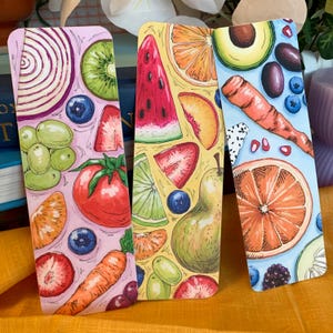 May include: Three colorful bookmarks featuring a variety of fruits and vegetables. The bookmarks are printed on a white background with a pink, yellow, and blue background. The bookmarks are approximately 6 inches long.