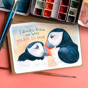 May include: A watercolor illustration of two puffins with the text "I always knew we were meant to beak". The puffins are facing each other with their beaks touching. The background is a soft yellow with orange and pink hues.