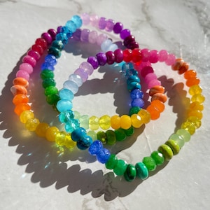 May include: Three rainbow colored beaded bracelets with different colored beads. The bracelets are made of various gemstones and are stacked on top of each other.