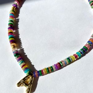 Pode incluir: Um colar de contas coloridas com um pingente de ouro em forma de letra A. As contas são feitas de várias pedras em tons de rosa, verde, azul, amarelo e vermelho.