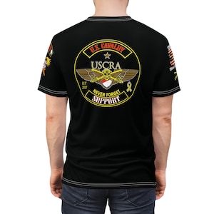Puede incluir: Camiseta negra con un gráfico circular amarillo que presenta una calavera y huesos cruzados, alas y el texto "USCRA, U.S. Cavalry, Est 2007, Never Forget, Support".