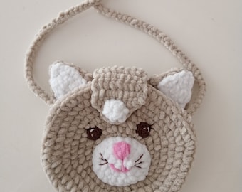 May include: A beige crocheted cat-shaped purse with a white face, pink nose, and brown eyes. The purse has a white strap.