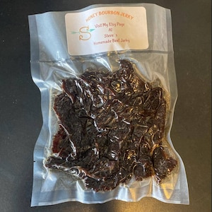 May include: A clear plastic bag filled with dark brown beef jerky. The bag has a label that reads "Honey Bourbon Jerky" and "Visit My Etsy Page At Steve's Homemade Beef Jerky".