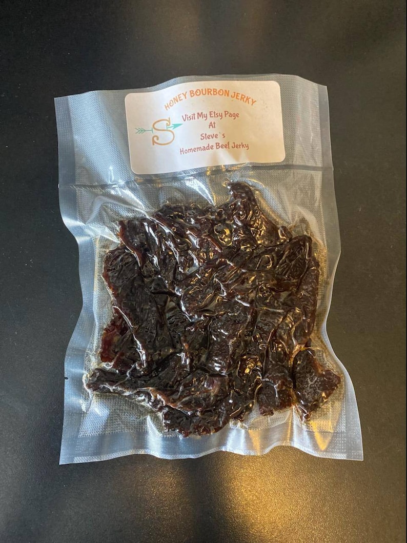 May include: A clear plastic bag filled with dark brown beef jerky. The bag has a label that reads "Honey Bourbon Jerky" and "Visit My Etsy Page At Steve's Homemade Beef Jerky".