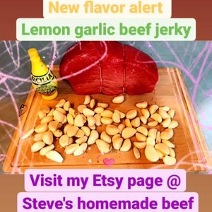 May include: A close-up of a wooden cutting board with a red beef roast, a bottle of lemon juice, and a pile of garlic cloves. The text "New flavor alert" and "Lemon garlic beef jerky" is displayed on the image. The text "Visit my Etsy page @ Steve's homemade beef yerky" is also displayed on the image.