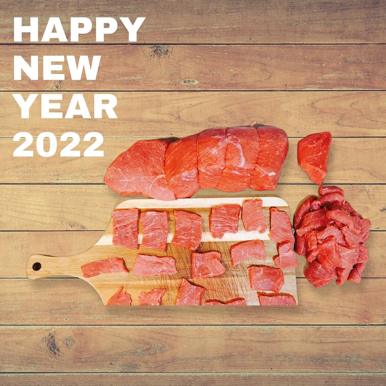 May include: Happy New Year 2022. A wooden cutting board with raw red meat, including a large piece of beef and smaller pieces of beef, arranged on the board.