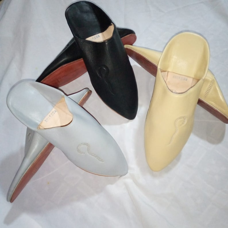 May include: Three leather babouche slippers in black, light gray, and cream. Each slip-on shoe features a pointed toe, a flat brown sole, and a small embroidered detail on the upper. The interior of each slipper is lined in a light tan color.