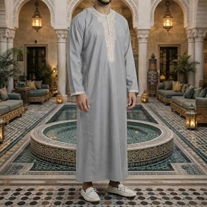 May include: A grey, long-sleeved robe with white trim and detailed embroidery around the neckline. The robe is worn with white loafers. The background shows an ornate architectural setting with a water feature.