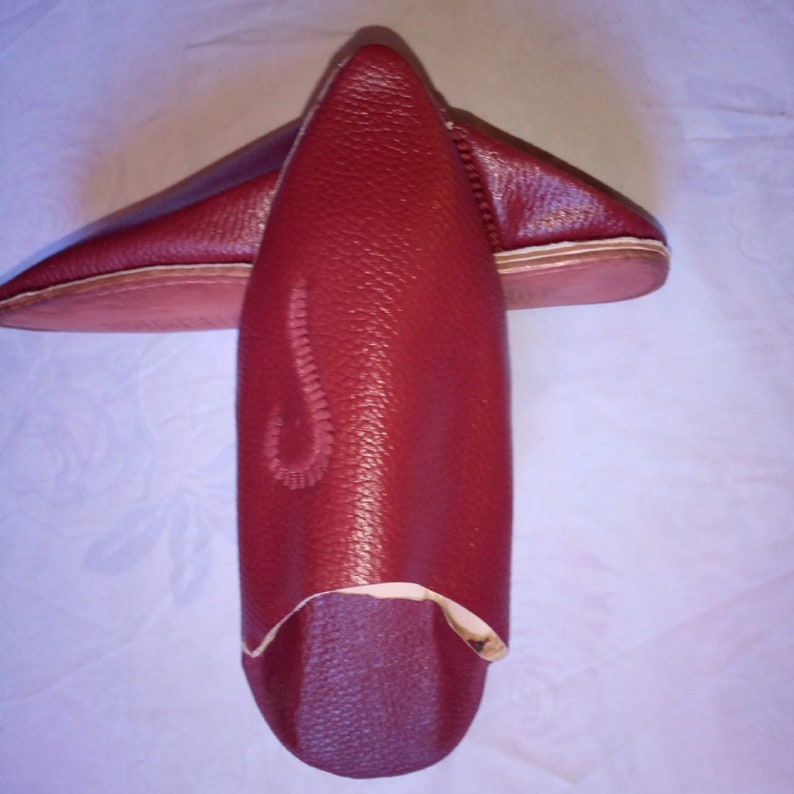 May include: A pair of deep red leather slippers with a pointed toe design. The slippers feature a textured surface and a decorative stitched pattern on the upper part of the shoe. The soles are flat and light-colored, contrasting with the dark red leather.