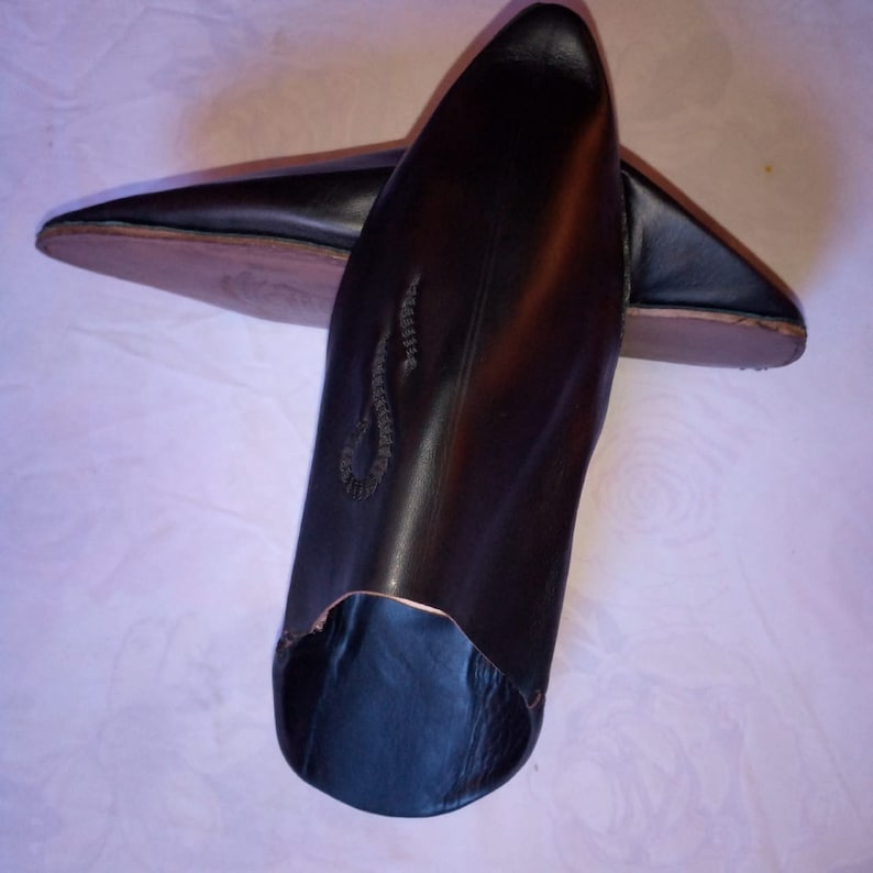 May include: A pair of black leather pointed-toe slippers featuring a smooth finish and a subtle, dark embroidered detail on the upper surface. The footwear has a low-cut opening and a tan-colored sole visible on the underside of the heel area.