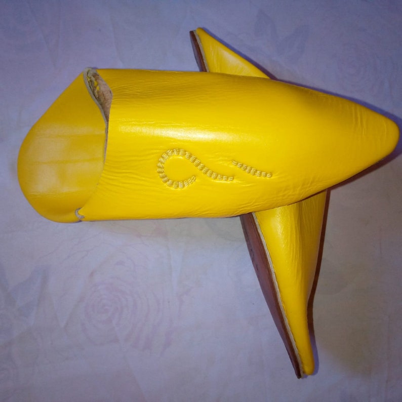 May include: A pair of bright yellow leather slippers featuring a pointed toe design and a subtle, curved decorative stitch pattern on the upper surface. The slippers have a smooth texture and a distinct, elongated shape with a flat brown sole.