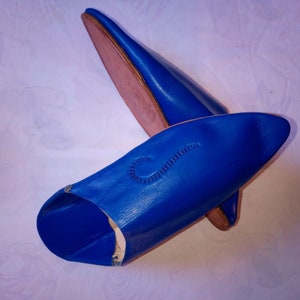 May include: A pair of vibrant blue leather slippers with a pointed toe design. The slippers feature a smooth finish on the exterior and a tan-colored interior lining. A decorative stitched detail is visible on the top surface of the front slipper.