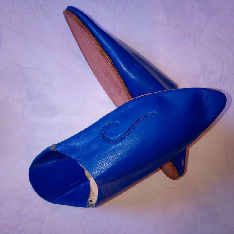 May include: A pair of vibrant blue leather slippers with a pointed toe design. The slippers feature a smooth finish on the exterior and a tan-colored interior lining. A decorative stitched detail is visible on the top surface of the front slipper.
