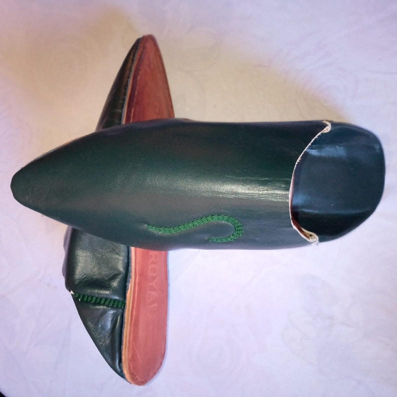 May include: A pair of dark green leather slippers with pointed toes and a low-cut opening. The slippers feature a decorative green stitched detail on the upper surface and a reddish-brown inner sole with the word ROYAL embossed on the heel.