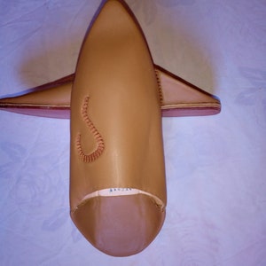 May include: A pair of tan leather babouche slippers with a pointed toe and a small, decorative embroidered S-shaped detail on the upper. The slippers feature a smooth finish, a low-profile design, and a visible white interior lining at the heel.