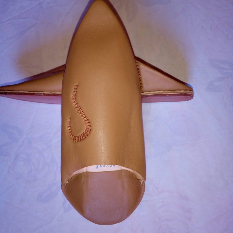 May include: A pair of tan leather babouche slippers with a pointed toe and a small, decorative embroidered S-shaped detail on the upper. The slippers feature a smooth finish, a low-profile design, and a visible white interior lining at the heel.
