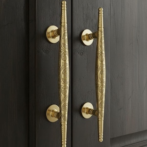 May include: Two brass door handles with intricate scrollwork and a warm, golden finish. The handles are mounted on a dark wooden door, showcasing their decorative design and adding a touch of elegance to the door's aesthetic.