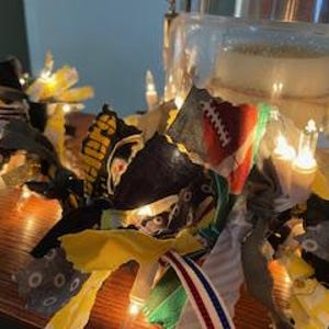 May include: A string of fabric scraps with a variety of colors and patterns, including yellow, black, green, and red, is strung with white fairy lights. The fabric scraps are tied together with knots and the lights are strung through the knots.