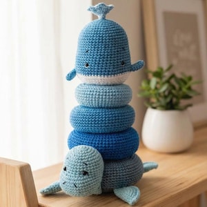 May include: A handmade crochet stacking toy in shades of blue, featuring a whale at the top and a turtle at the base. The toy includes several stacking rings in varying shades of blue. The toy is sitting on a wooden surface.