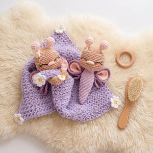 May include: Crocheted baby toys and accessories. The image features two handmade, light brown and pink crocheted dolls with flower accents, a lavender blanket, a wooden teething ring, and a wooden brush, all arranged on a cream-colored, textured surface.