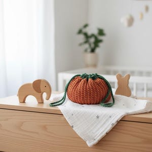 Op de afbeelding: Een oranje gehaakte trekkoordtas met groene banden ligt op een witte stof met sterrenpatroon. Een houten olifant en een houten konijn staan op een licht houten oppervlak. De scène suggereert een kinderkamer.