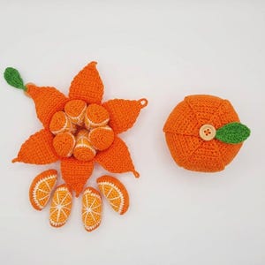 May include: Crocheted orange fruit set, featuring a large orange flower with segments and a whole orange with a button and leaf. The set includes orange slices and small orange segments. The colors are orange, white, and green.