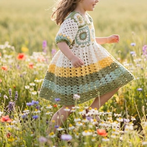 May include: A handmade crochet dress in a field of wildflowers. The dress features a square yoke and short sleeves, with a skirt of alternating stripes of cream, yellow, green, and blue. The dress is knee-length and appears to be worn by a child.