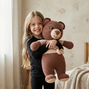 Puede incluir: Un gran oso de peluche de ganchillo hecho a mano en tonos marrón y beige. El oso tiene una cabeza redonda, orejas pequeñas y una cara sonriente. El oso está siendo abrazado por una persona que lleva una camiseta negra de manga larga.