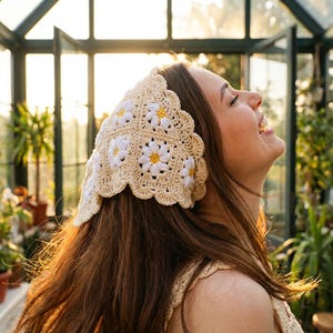 May include: A cream-colored crochet headband featuring white and yellow daisy flower designs. The headband has a scalloped edge and is worn on the head. The image is taken in a greenhouse with sunlight.
