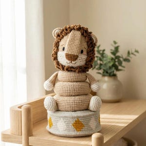 Puede incluir: Un juguete de león de ganchillo hecho a mano en beige, marrón y blanco. El león tiene una cabeza redonda con una melena marrón, una cara blanca y brazos y piernas pequeños. Se asienta sobre una pila de anillos sobre una base gris y amarilla.