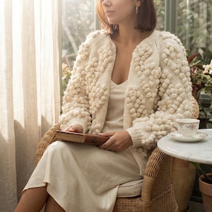 May include: A cream-coloured, open-front cardigan with a textured bobble-stitch design. The cosy sweater is worn over a cream-coloured dress. A book and teacup are visible. The scene is set in a sunlit room with a wicker chair.