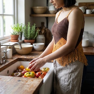 May include: A brown and orange crochet top with fringe detail. The top is paired with linen pants. A variety of colorful tomatoes are in a white sink. The kitchen has wooden shelves and a window.