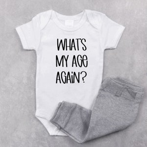 May include: White baby onesie with black text that reads "WHAT'S MY AGE AGAIN?" and a pair of gray sweatpants.