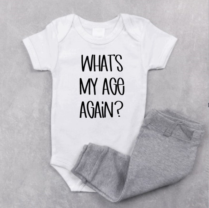 May include: White baby onesie with black text that reads "WHAT'S MY AGE AGAIN?" and a pair of gray sweatpants.
