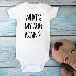 May include: White baby onesie with black text that reads "What's my age again?"  The onesie is lying on a light blue wooden surface with a brown stuffed animal.