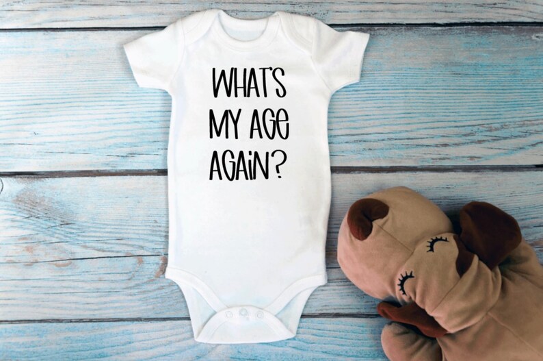 May include: White baby onesie with black text that reads "What's my age again?"  The onesie is lying on a light blue wooden surface with a brown stuffed animal.