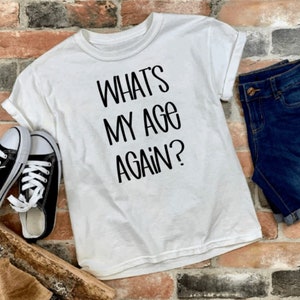 May include: White t-shirt with black text that reads "What's My Age Again?" The shirt is laid out on a brick surface with a pair of black sneakers and blue denim shorts.
