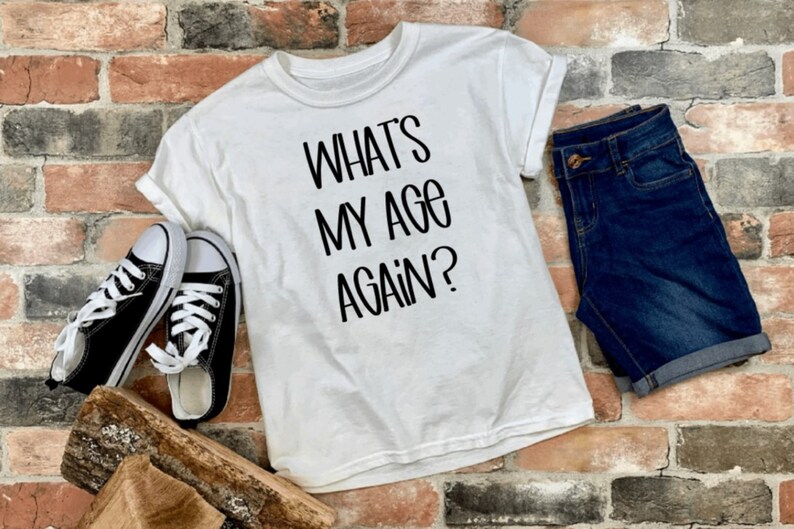 May include: White t-shirt with black text that reads "What's My Age Again?" The shirt is laid out on a brick surface with a pair of black sneakers and blue denim shorts.