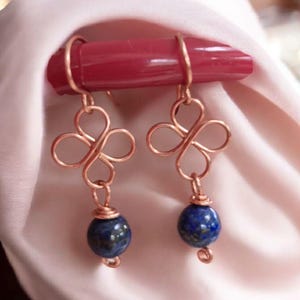以下が含まれることがあります： 四つ葉のクローバーのデザインと濃い青色の球形ビーズが付いた銅線イヤリング。 イヤリングは赤い円筒形の物体から吊り下げられています。 背景は柔らかいピンク色の生地です。 長さ約2cm。