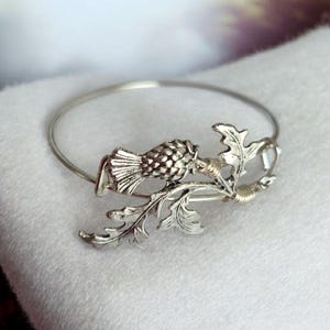 Può includere: Bracciale rigido argentato con un elemento decorativo dettagliato. Il punto focale è un design stilizzato di pesce o uccello, completato da intricati accenti di foglie e rami. Il bracciale è posto su uno sfondo bianco morbido.