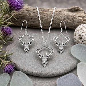 May include: A silver-toned jewelry set consisting of a necklace and a pair of drop earrings. Each piece features a detailed deer head pendant with prominent antlers, suspended from a delicate silver chain and standard earring hooks.