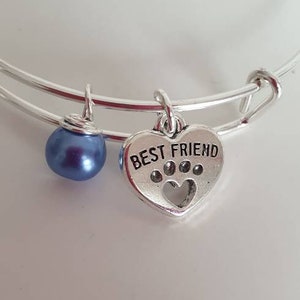 Könnte beinhalten: Silbernes, ausziehbares Armband mit einem blauen Perlenanhänger und einem Herzanhänger mit der Aufschrift "Bester Freund" und einem Pfotenabdruck im Inneren.
