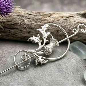 May include: A silver-toned metal hair pin featuring a detailed thistle motif with spiky leaves and a textured flower head. The design includes a circular frame and a long, slender pin with a decorative curled end, crafted from polished metal.
