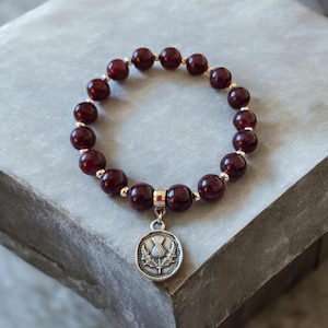 Puede incluir: Una pulsera con cuentas redondas de color rojo oscuro y detalles plateados. Un dije plateado con un diseño de cardo cuelga de la pulsera. La pulsera se muestra sobre una superficie gris.